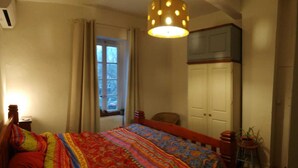 A bedroom with a bed, a window with curtains, a door, a potted plant, and a hanging light fixture.