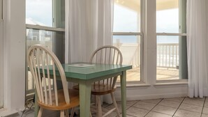 A room with a table and two chairs, large windows with white curtains, and a view of a wooden deck. - Simple Pleasures, Gulf Beachfront, 3 Bedroom, 2 Bath (Dauphin Island)