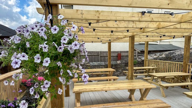 An outdoor wooden deck with picnic tables and a pergola with string lights. - The George Inn Plympton (Plymouth)