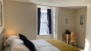 A bedroom with a bed, a window with curtains, a chair, and a small table with a lamp. - The George Inn Plympton (Plymouth)