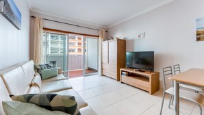 A modern living room with a sofa, TV, and dining area, featuring a balcony with a view of buildings. - Bright 1 BDR Flat W/ Pool & Balcony by LovelyStay (Portimão)
