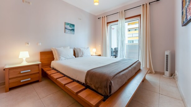 A bedroom with a bed, bedside tables, a lamp, a painting, a window with curtains, and a radiator. - Bright 1 BDR Flat W/ Pool & Balcony by LovelyStay (Portimão)