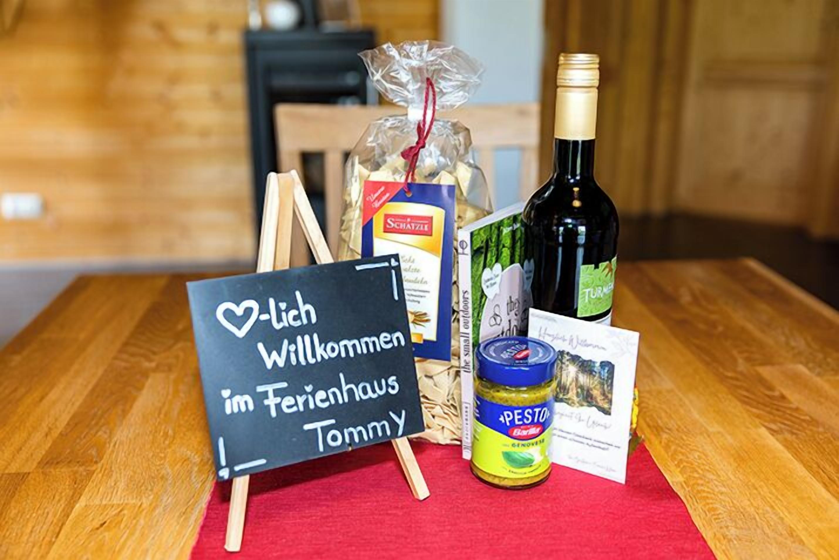 A welcome sign, a bottle of wine, a jar of pesto, a box of pasta, and a bag of snacks on a table.