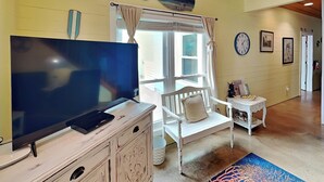 Un salon chaleureux avec un téléviseur posé sur une commode, un fauteuil blanc, une petite table avec une lampe et une grande fenêtre avec des rideaux.