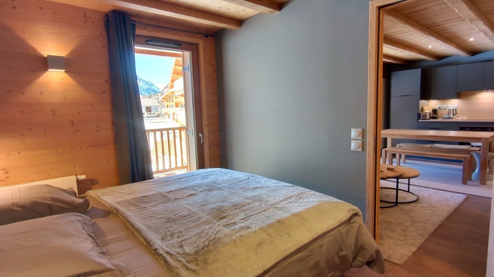 A bedroom with a bed, a wooden ceiling, a window with a view of buildings and mountains, and a kitchenette area with a table and chairs.