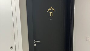 Eine schwarze Tür mit Bergmotiv und der Zahl 11, eine Fußmatte mit Hirschkopf und eine weiße Wand mit Lichtschalter.