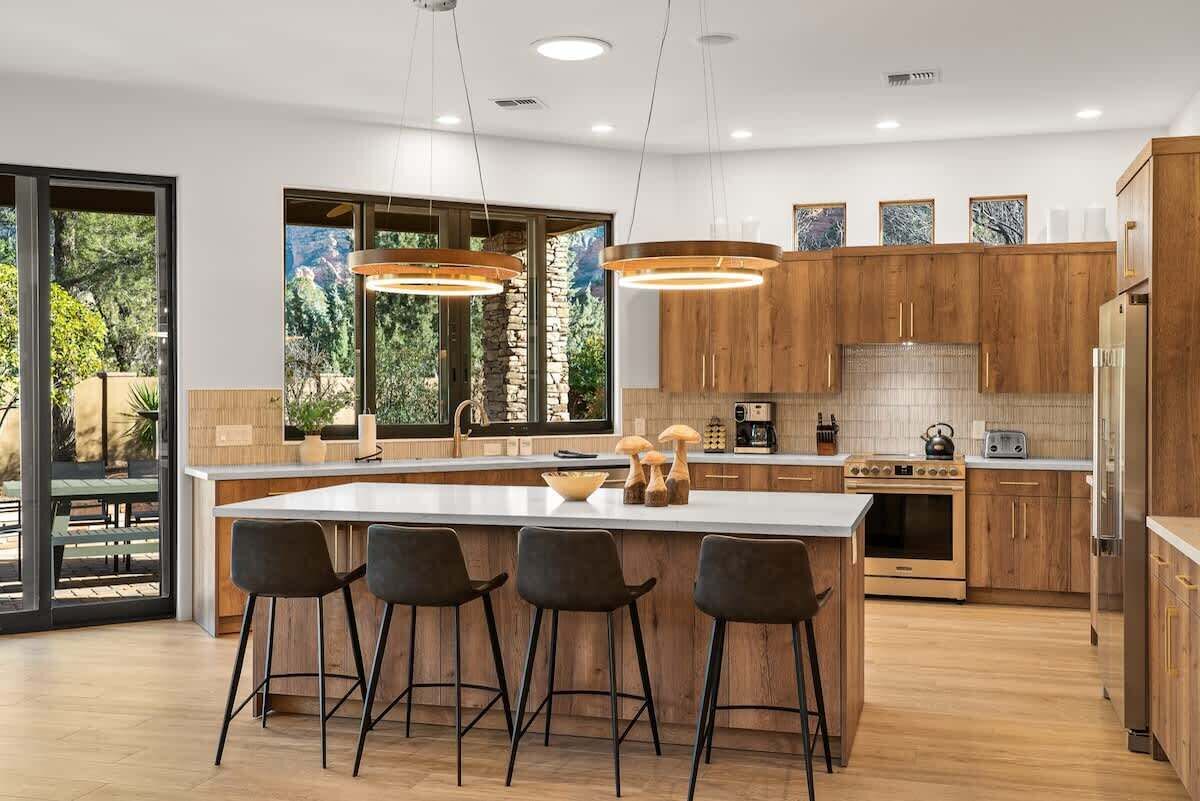 A modern kitchen with wooden cabinets, a central island with bar stools, and a large window offering a view of the outdoors.