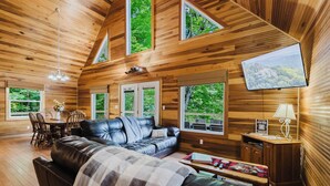 A cozy cabin interior with a leather sofa, dining table, and large windows offering views of the forest.