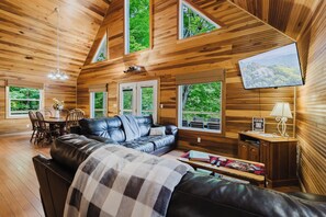A cozy cabin interior with a leather sofa, dining table, and large windows offering views of the forest.