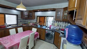 A kitchen with wooden cabinets, a washing machine, a large water dispenser, and a dining area with a pink tablecloth. - Modern Duplex In the center of Irbid (Irbid)