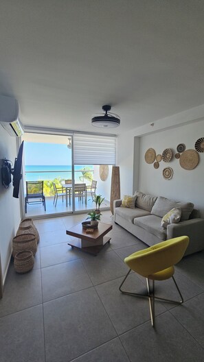 A living room with a sofa, a yellow chair, a coffee table, and a view of the ocean through a sliding door.