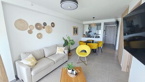 Un salon avec un canapé, une table basse et des fauteuils jaunes. On y trouve également un coin repas avec une table et des chaises, et une cuisine à l’arrière-plan.