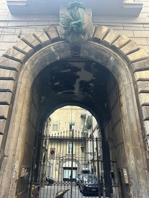 Un portail en pierre en arc de cercle, surmonté d’une grille métallique, mène à un bâtiment à la façade jaune devant lequel est garée une voiture noire.