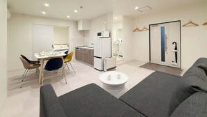 A modern living room with a sofa, a dining table with chairs, a kitchen area with appliances, and a door with a coat rack.