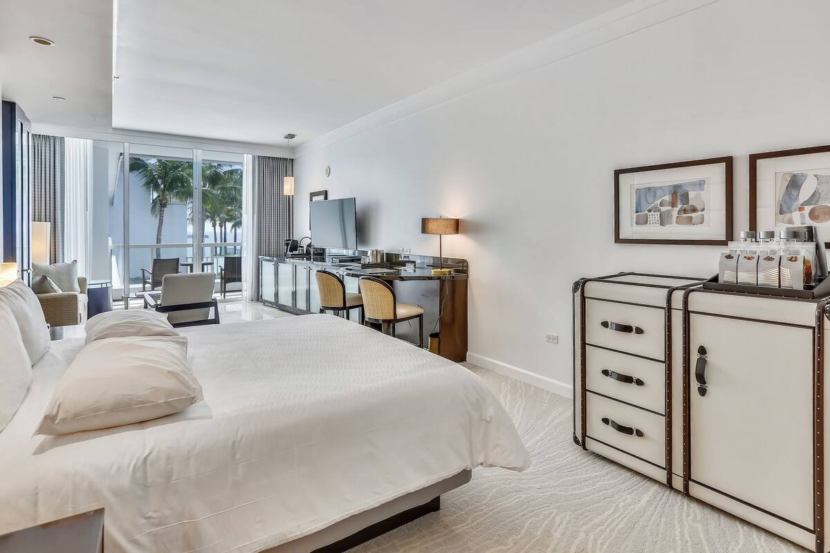 A modern hotel room with a large bed, a desk, and a view of palm trees through large windows.