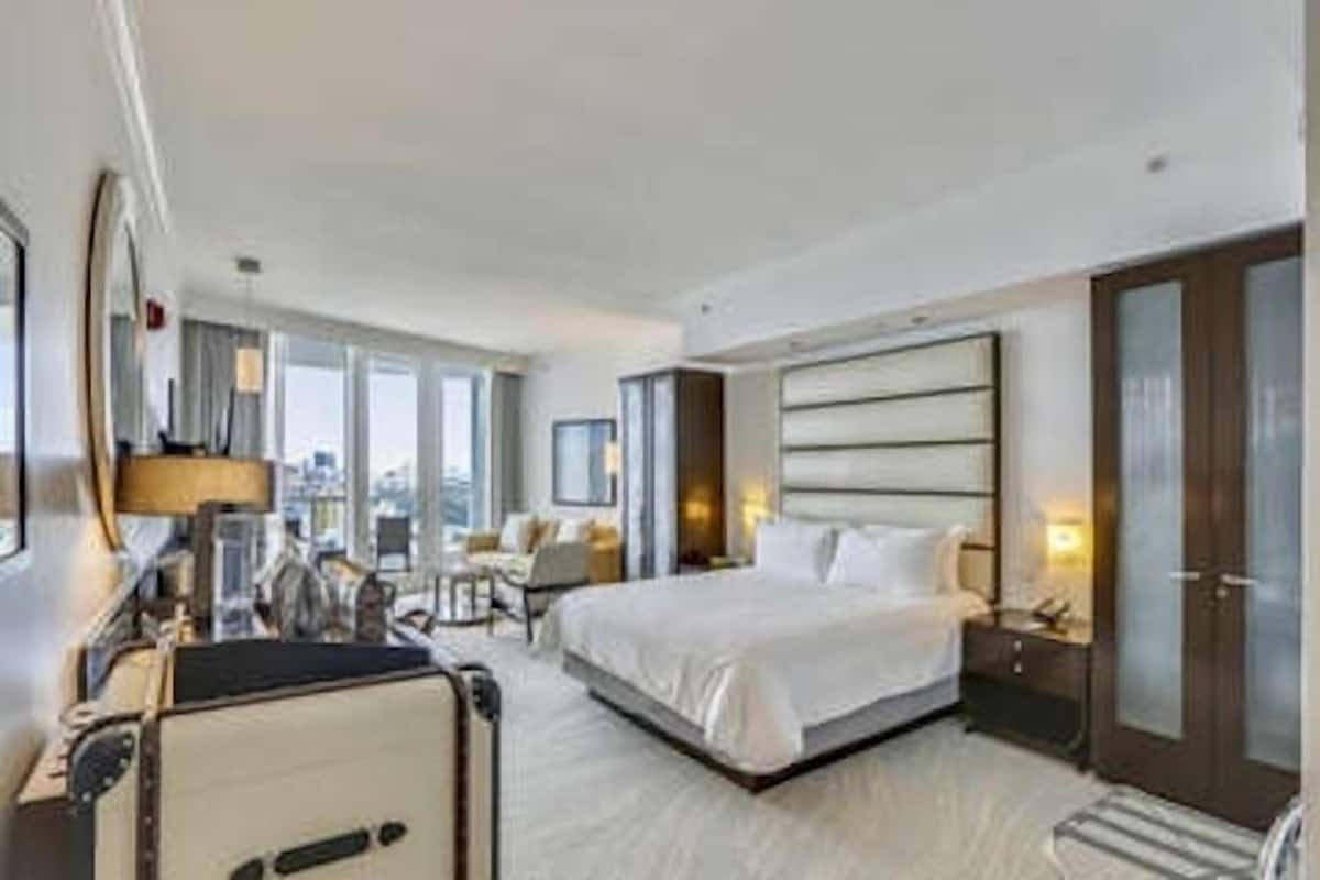 A hotel room with a large bed, a desk with a chair, a wardrobe, and a view of the city.