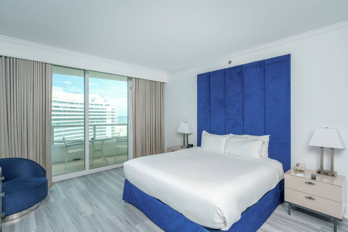 A hotel room with a large bed, a blue accent wall, a glass door leading to a balcony, and a blue chair.