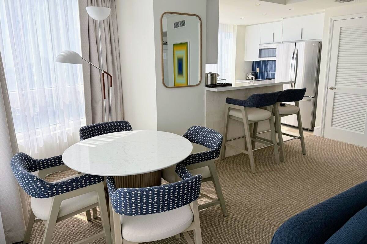 A modern hotel room with a round dining table, four patterned chairs, a kitchenette, and a large window with curtains.