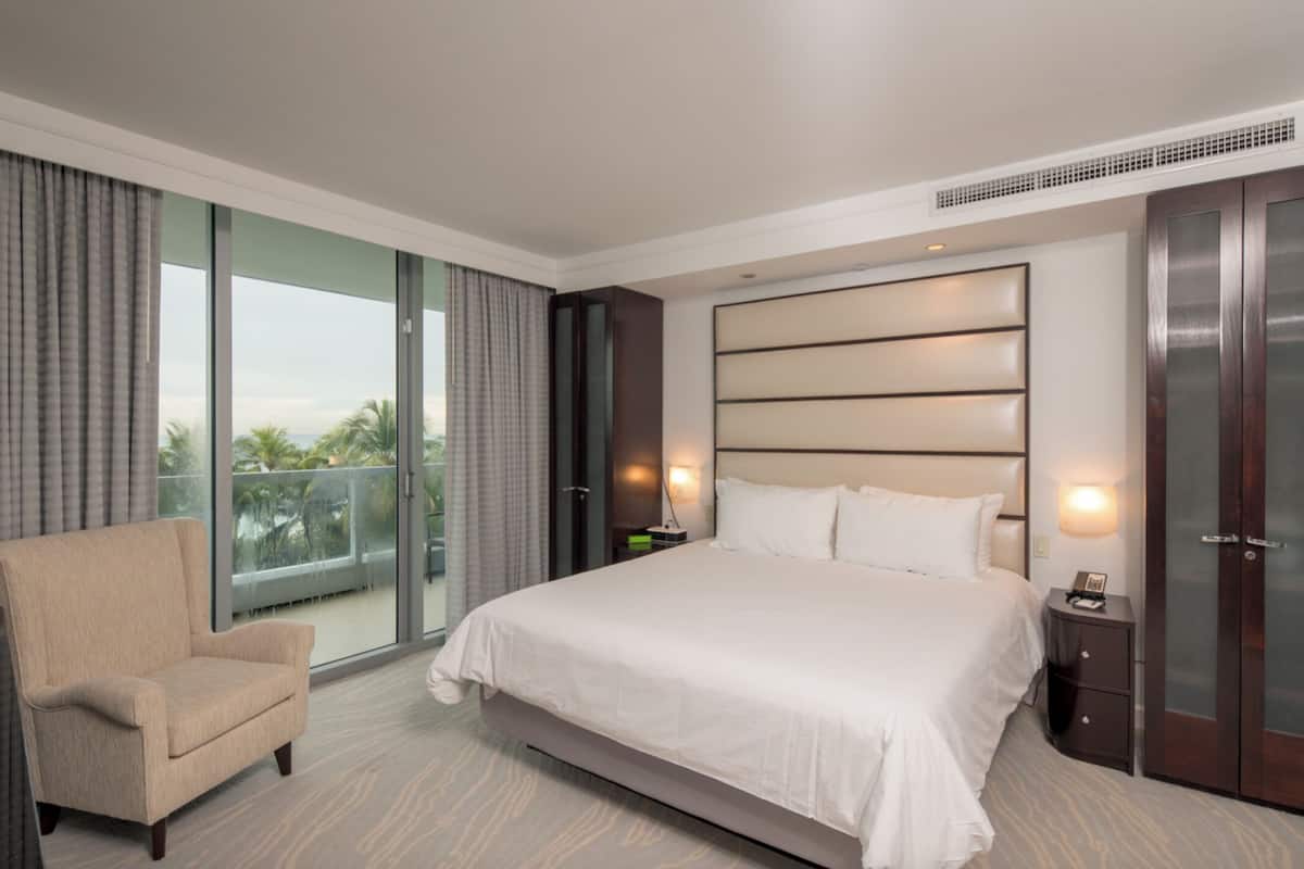 A modern hotel room with a large bed, a chair, a nightstand, and a view of the outdoors.