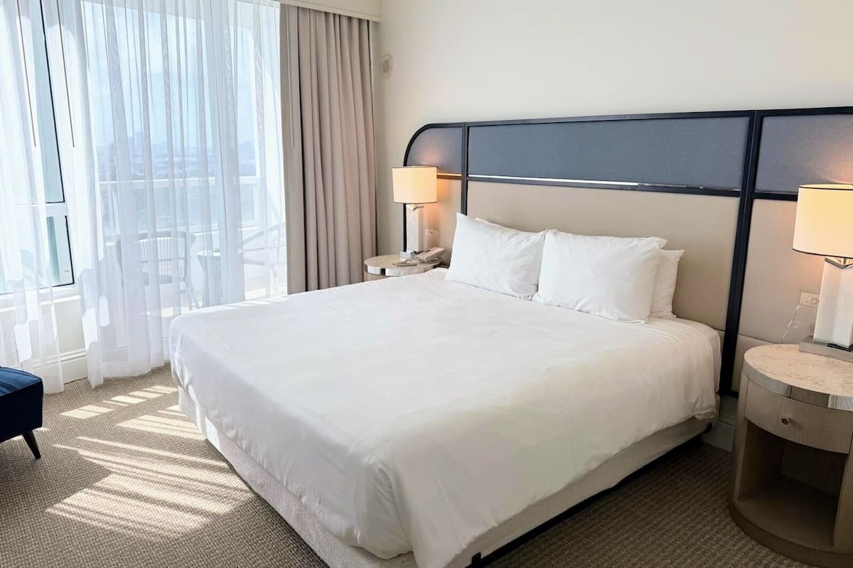 A hotel room with a large bed, two bedside tables with lamps, a view of the city through sheer curtains, and a balcony with outdoor furniture.