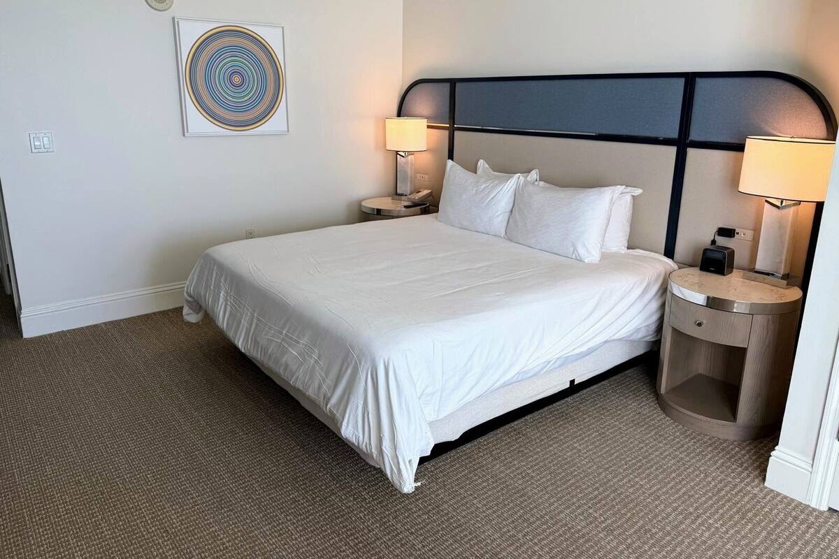 A hotel room with a large bed, two bedside tables with lamps, a wall clock, and a framed artwork.