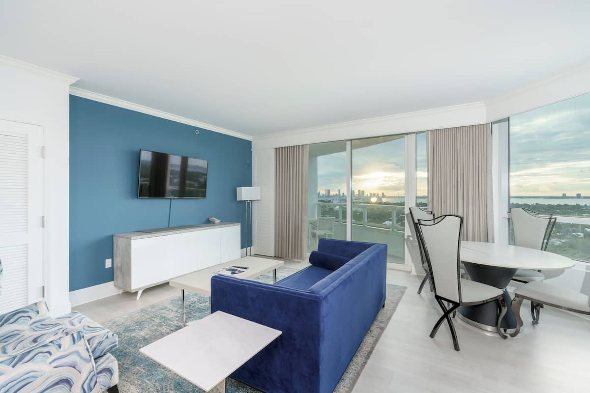 A modern living room with a blue sofa, a white coffee table, a TV mounted on the wall, and a dining area with a table and chairs.