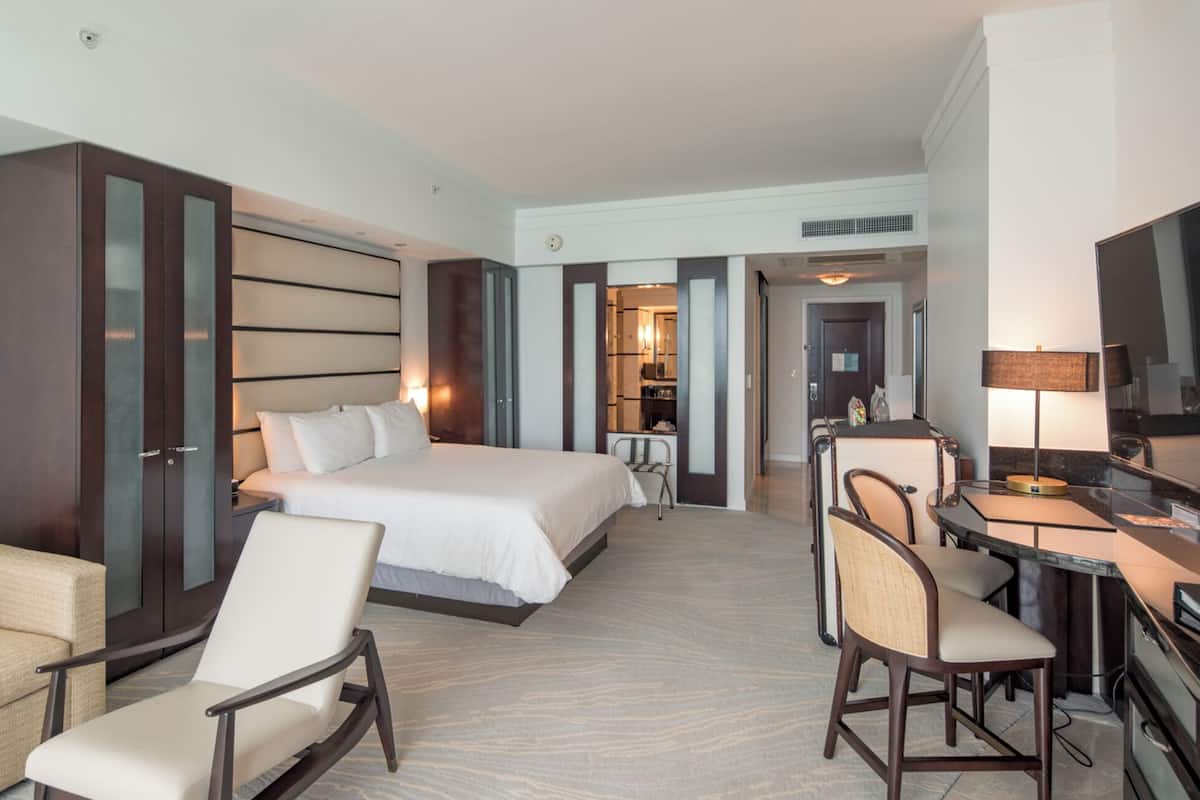 A hotel room with a large bed, two chairs, a desk with a lamp, and a television.
