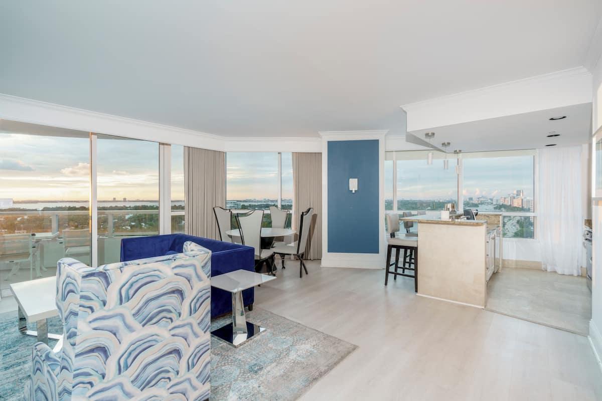A spacious living area with a large window, a blue sofa, a dining table, and a kitchen area.
