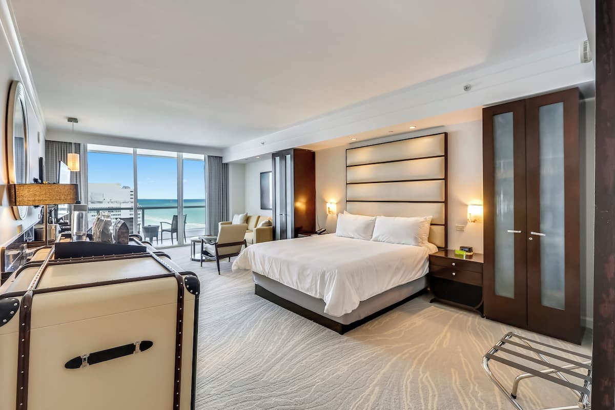A modern hotel room with a large bed, a desk, a chair, and a view of the ocean.