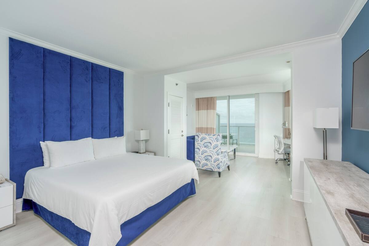A modern hotel room with a large bed, a desk, and a view of the balcony.