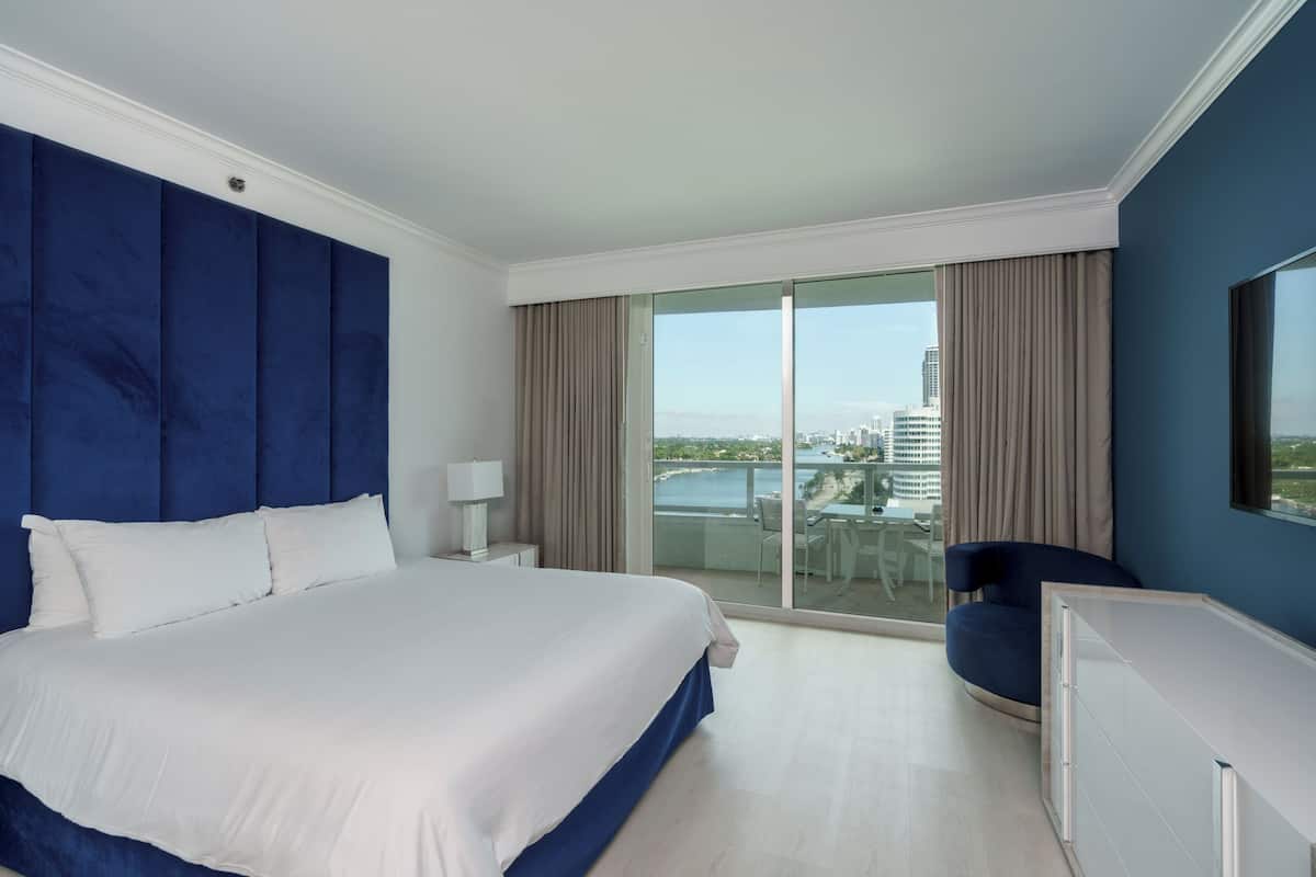 A hotel room with a large bed, a TV, a balcony with a view of the city, and a chair.