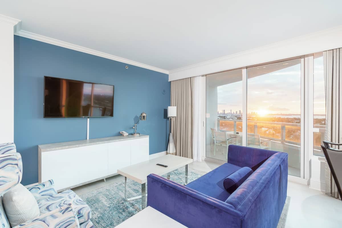 A modern living room with a blue wall, a flat-screen TV, a sofa, a coffee table, and a view of a cityscape through large windows.