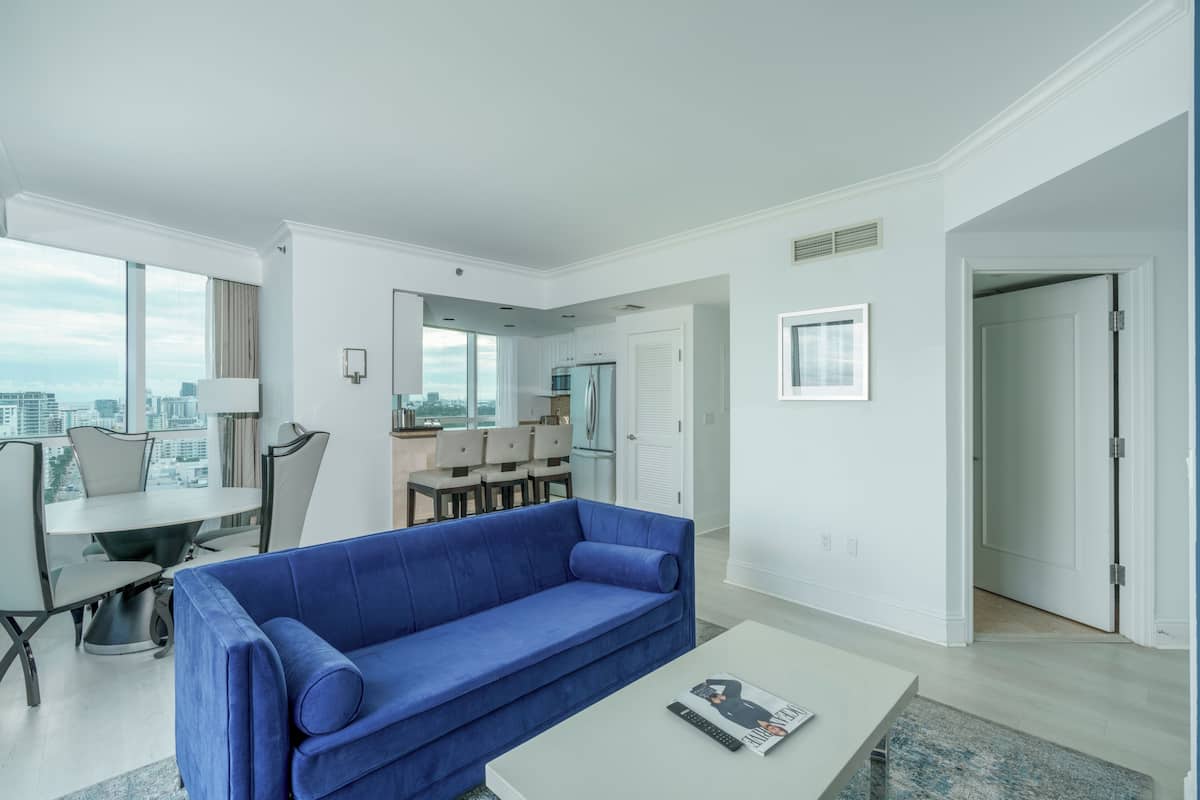 A modern living room with a blue sofa, a dining table, and a kitchen area visible through an open door.
