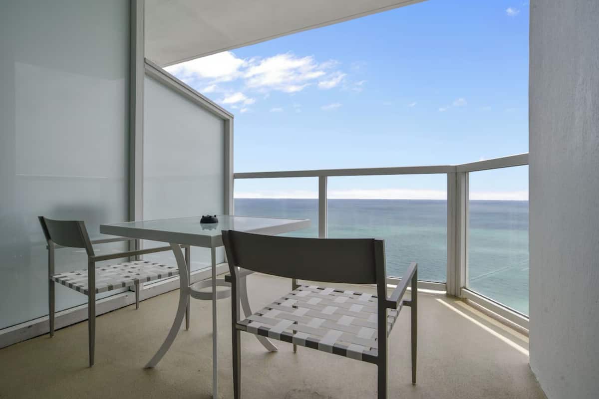 A balcony with a glass table and chairs, overlooking a body of water.