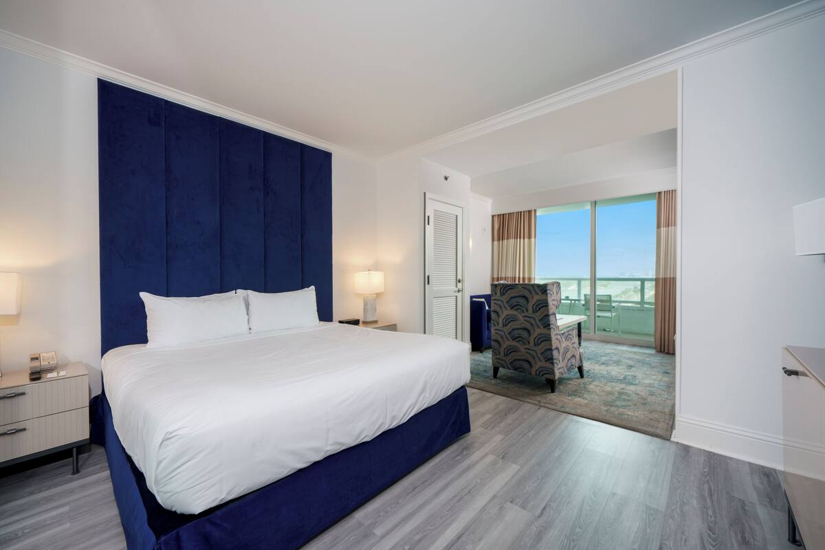 A modern hotel room with a large bed, a nightstand, a chair, and a view of the outdoors.