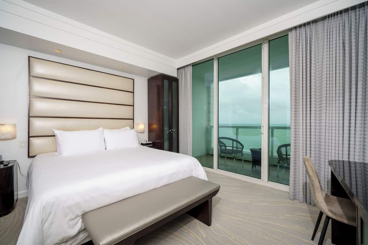 A hotel room with a large bed, a bench, a desk, and a balcony with chairs.