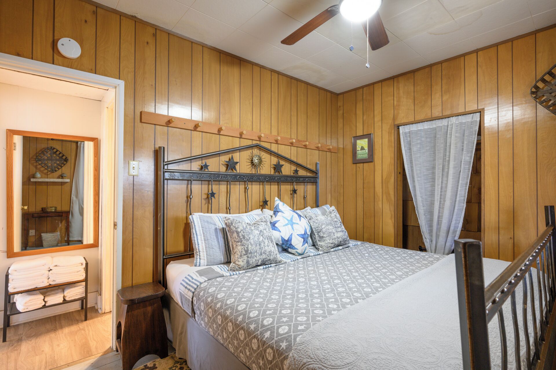 Une chambre avec un mur lambrissé en bois, un lit avec une literie à motifs, une table de chevet, une commode, un ventilateur de plafond et une fenêtre avec un rideau.