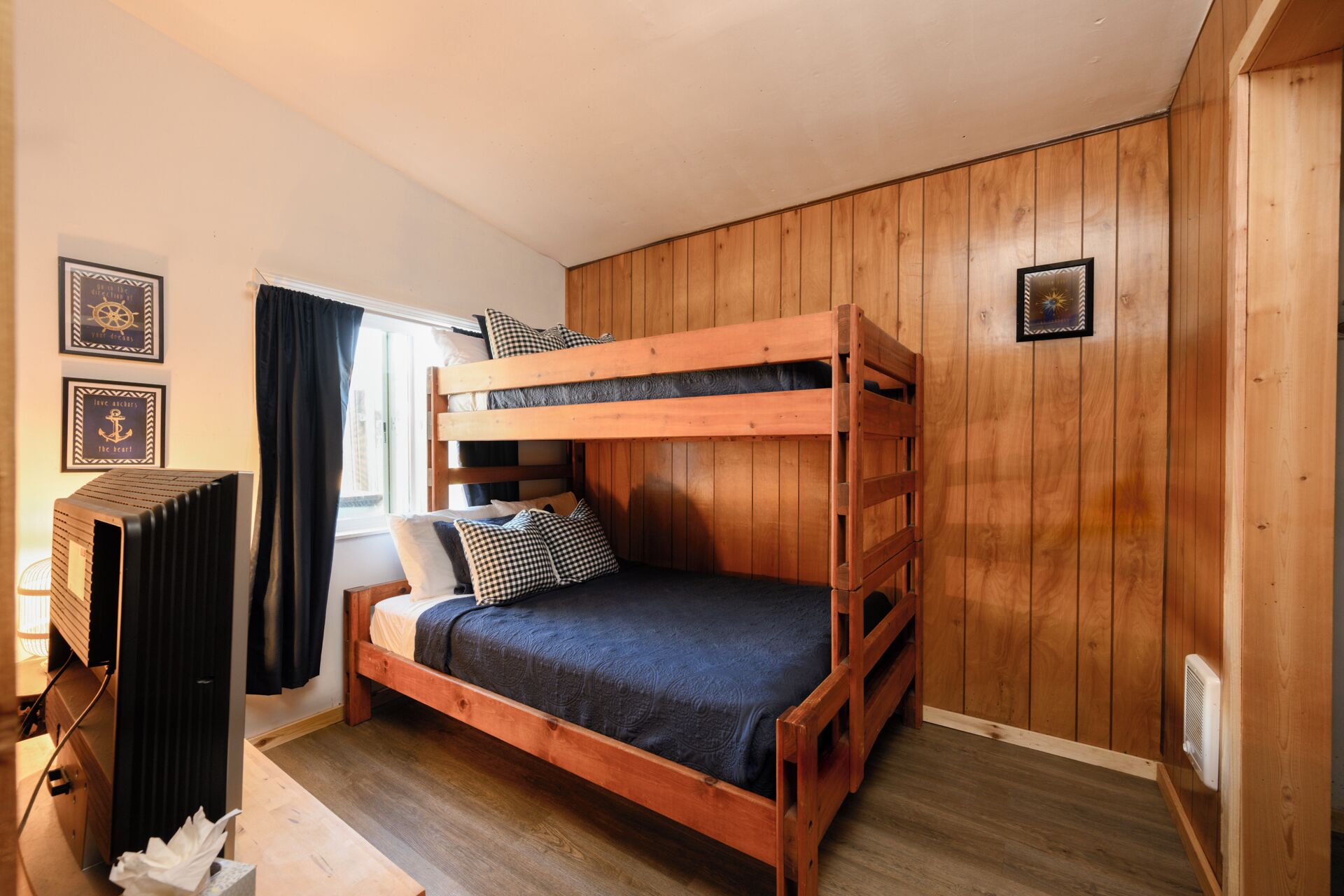 A bunk bed room with wooden walls, a window with curtains, and framed pictures on the wall.
