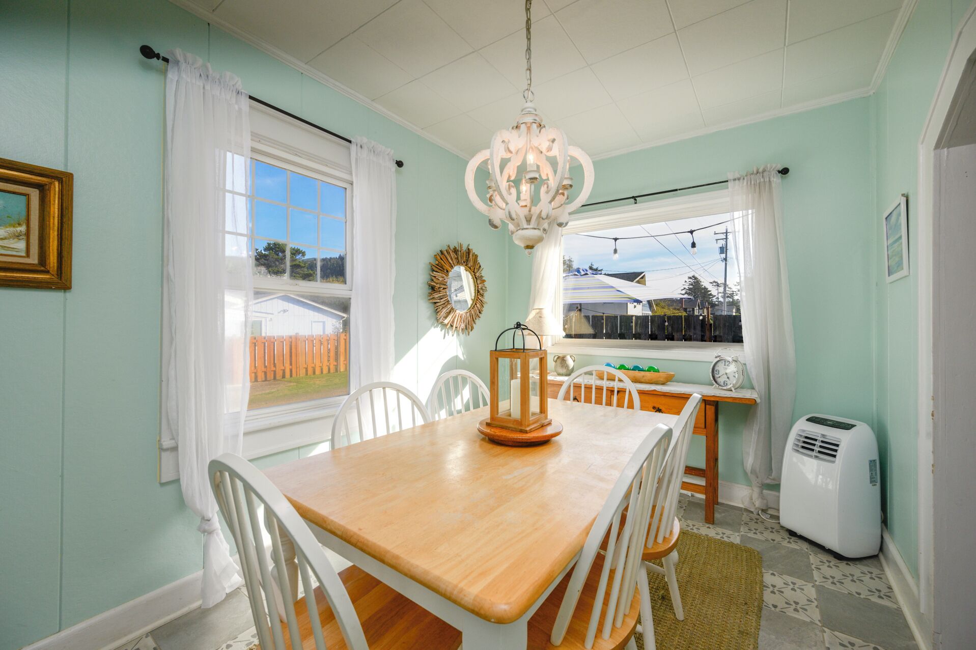 A dining room with a wooden table, white chairs, a chandelier, and a view of the outside.