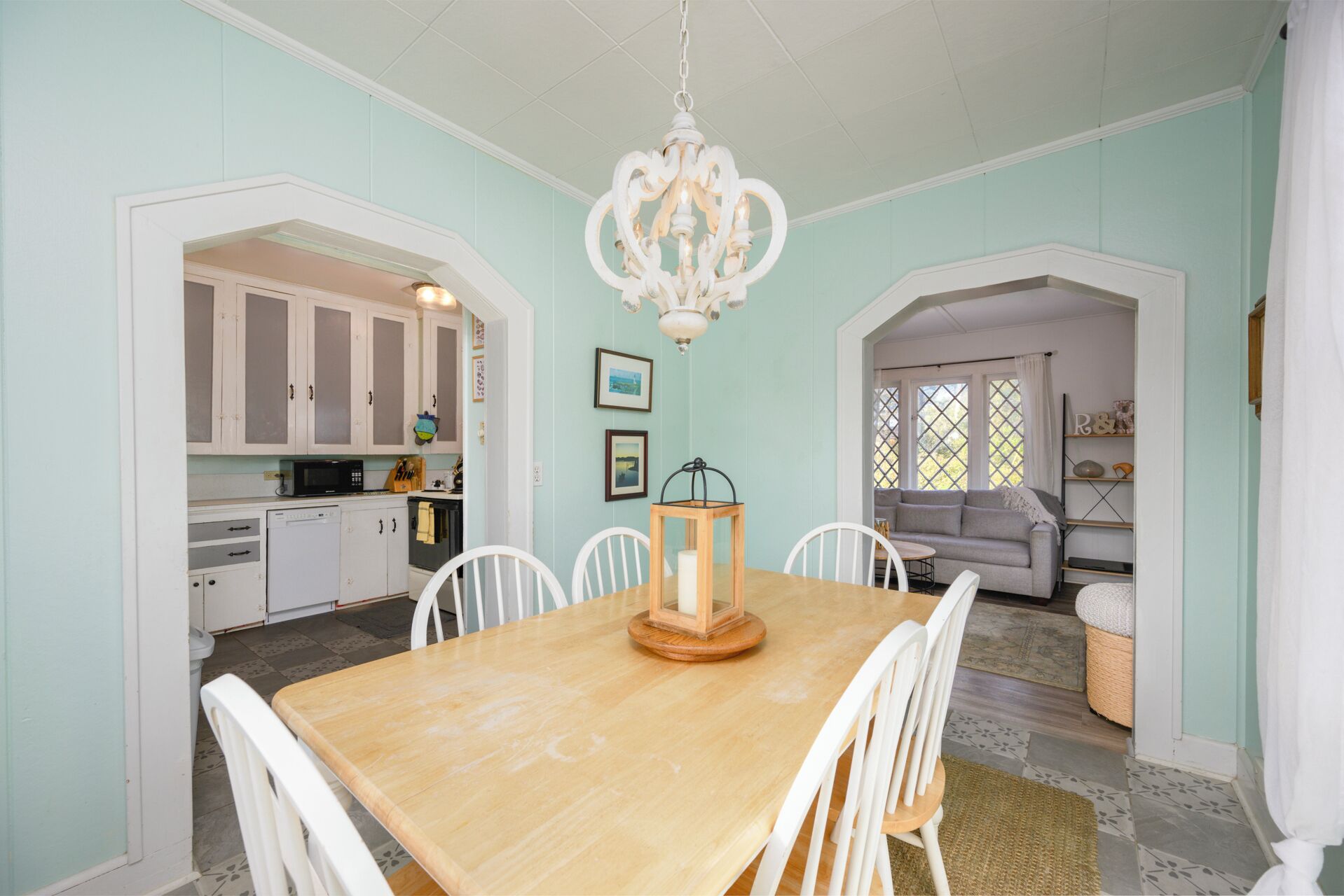A dining area with a wooden table and white chairs, a chandelier, and a kitchen in the background.