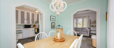 A dining area with a wooden table and white chairs, a chandelier, and a kitchen in the background.