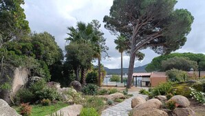 Un sentier de jardin bordé de gros rochers et de différentes plantes, qui mène à un bâtiment doté d’un toit en tuiles.