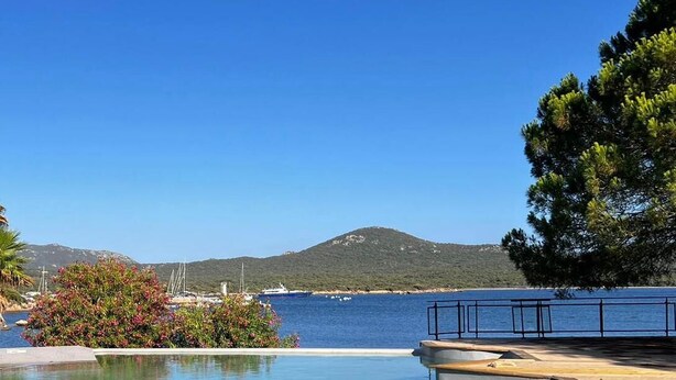 Un ciel d’un bleu limpide, une terrasse en bois et une piscine offrant une vue sur la mer et une montagne en arrière-plan.