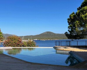 Un ciel d’un bleu limpide, une terrasse en bois et une piscine offrant une vue sur la mer et une montagne en arrière-plan.