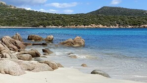 Une plage à l’eau d’un bleu limpide, bordée de gros rochers, avec une colline boisée en arrière-plan.