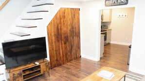 A modern living room with a wooden TV stand, a staircase with metal railings, and a kitchen area visible through an open door.
