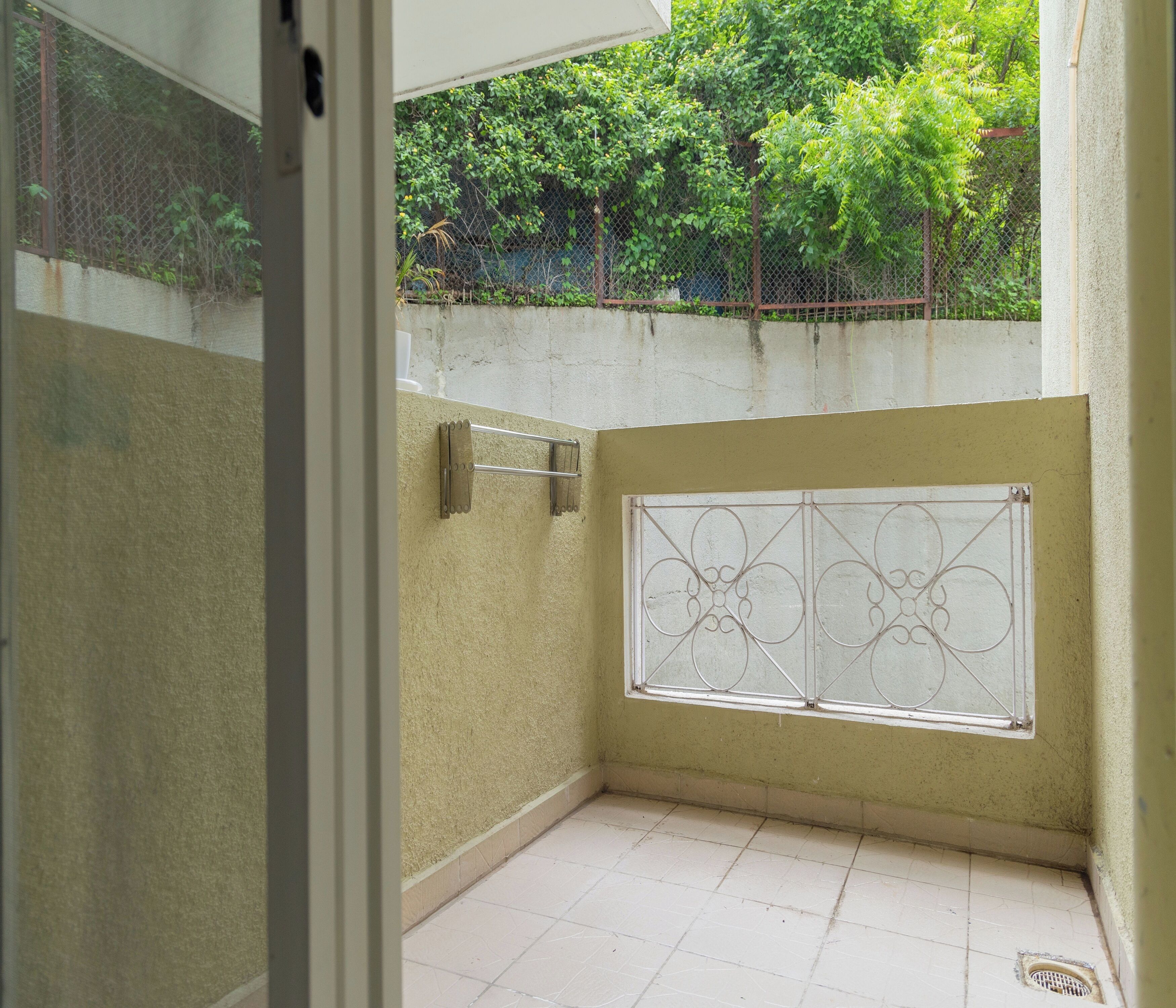 A small, enclosed balcony with a metal railing, a window with decorative bars, and a view of greenery beyond a fence.