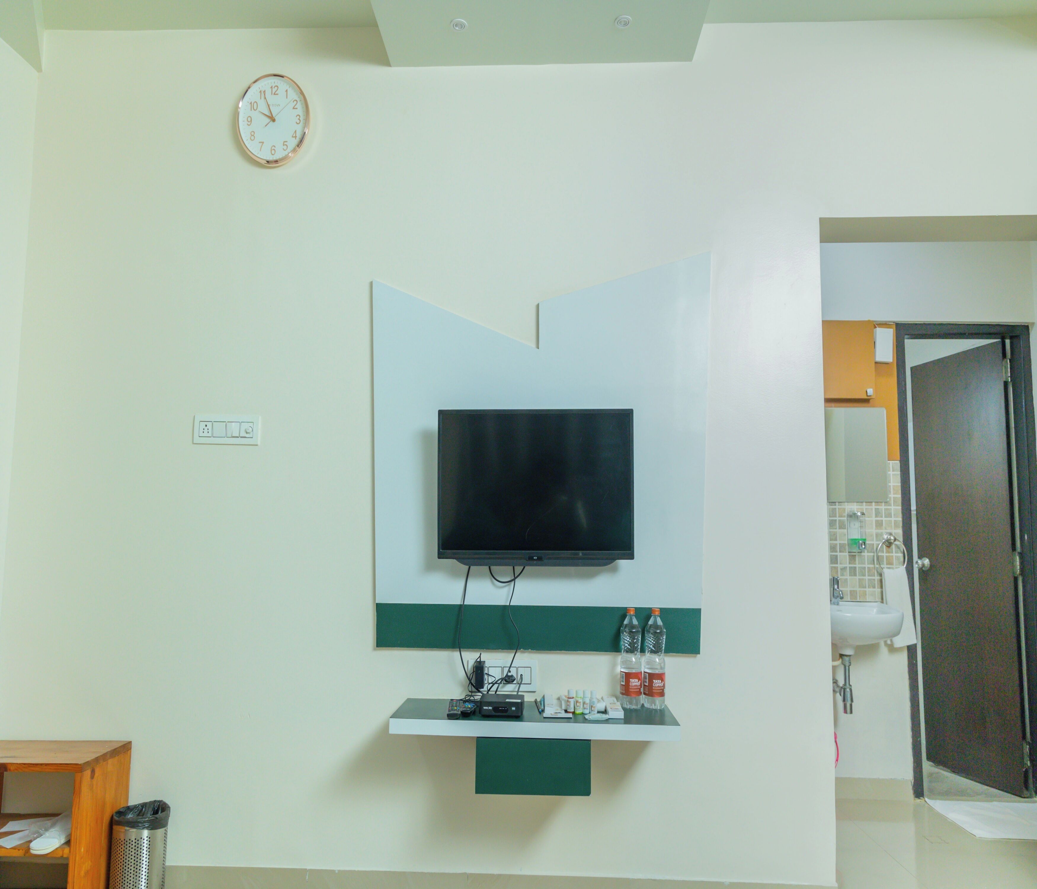 A room with a mounted TV, a shelf with electronic devices, and a clock on the wall.