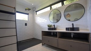 Une salle de bain moderne avec des murs en briques blanches, une grande fenêtre, deux miroirs ronds, un lavabo avec un receveur noir et un meuble-lavabo en bois.