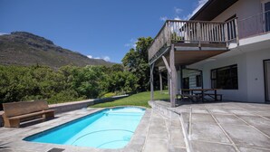 Une piscine avec un banc en bois, un patio couvert avec une table et des chaises, et une vue sur une montagne.